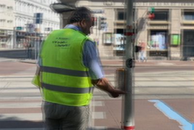Kreuzung in Wien, Gründerzeithäuser, Straßenbahngleise, Zebrastreifen. Ein Mann (mit gelber Warnweste) betätigt akustische Ampel mit Vibrationssignal