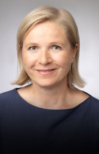 Portraitfoto: Eine Frau mit hellblonden, halblangen Haaren und dunkelblauem U-Boot-Shirt, grün-braunen Augen, lächelnd.