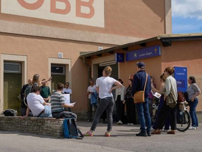 Eine Menschengruppe steht bzw. sitzt am Vorplatz vor einem orangen Gebäude (dem Bahnhofsgebäude) und hört einer Frau zu, die auf etwas deutet.