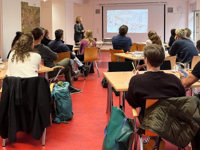 Vortrag mit Beamerpräsentation. Auditorium sitz an locker angeordneten Tischen