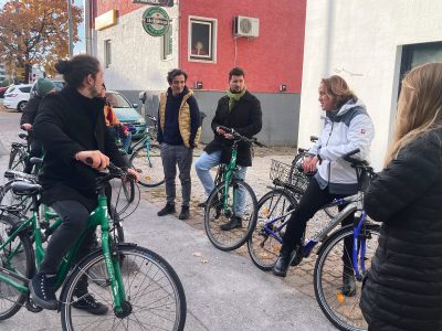 Gruppe von radfahrenden Personen steht im Halbkreis, eine Person trägt vor. Im Hintergrund kleines Haus mit roter Fassade mit einem Gastronomiebetrieb