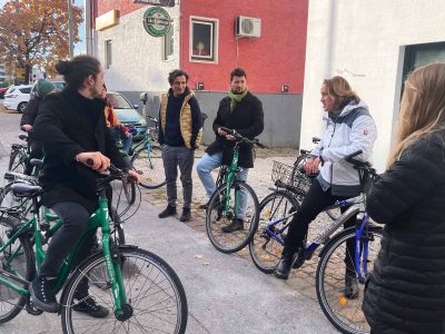 Gruppe von radfahrenden Personen steht im Halbkreis, eine Person trägt vor. Im Hintergrund kleines Haus mit roter Fassade mit einem Gastronomiebetrieb
