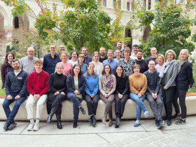 Gruppenfoto der Transformator:innen Teilnehmer:innen. Alle wirken sehr fröhlich und lachen herzlich. Der hintere Teil steht, die Vorderen sitzen auf einer langen Sitzbank aus Beton mit Holzbeplankung oben. Im Hintergrund viele Sträucher und Bäume.