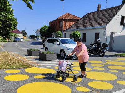Eine ältere Frau mit Rollator überquert die Straße auf einer Querungshilfe innerorts. Gelbe Kreisflächen unterschiedlicher Größe verbinden gegenüberliegende Gehsteige. Pflanztröge stehen am Fahrbahnrand und flankieren die Querungshilfe. Im Hintergrund einstöckige Häuser. Bäume säumen die Straße. Ein Auto hält, damit die Fußgängerin ungehindert queren kann.