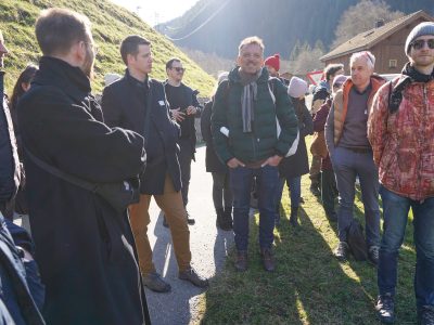 Gegenlichtfoto eine Personengruppe einer Führung auf Weg. Links und Rechts Wiese. Hinten Berge. Winterliche Bekleidung