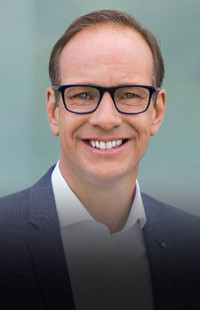 Portraitfoto: lächelnder Mann mit Brille mittleren Alters, Brille, weißes Hemd (offen getragen), blaues Sakko