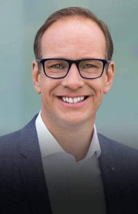 Portraitfoto: lächelnder Mann mit Brille mittleren Alters, Brille, weißes Hemd (offen getragen), blaues Sakko