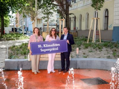 Drei Personen (Planungsstadträtin Ulli Sima, Bezirksvorsteher Marcus Franz sowie NEOS Wien Klubvorsitzende Selma Arapovic ) halten eine Schild "10., Klimafittes Supergrätzl". Im Hintergrund große Pflanzbeete mit Bestandsbäumen und zahlreichen neuen Jungbäumen. Im Vordergrund ein Wasserspiel